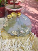 Handcrafted smokey grey resin pooja thali featuring golden leaf accents, pearl jewel centerpiece, Kundan peacocks, and three golden diya holders.