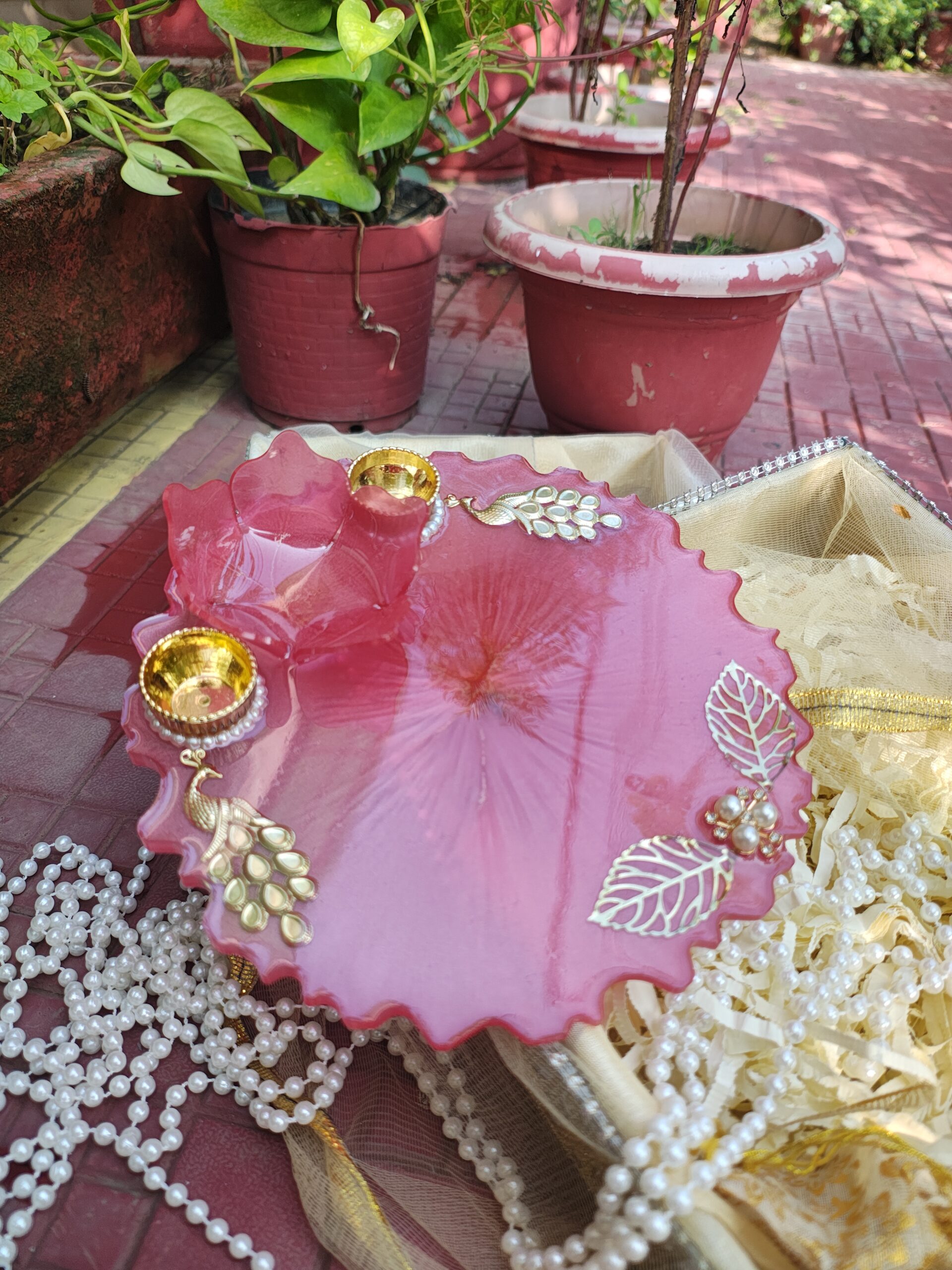 Handcrafted rose pink resin pooja thali featuring 3D lotus bowl, golden leaf filigree, Kundan peacock motifs, and pearl-trimmed diya holders.